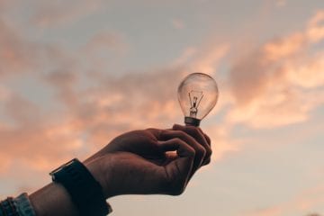 Person hält Glühbirne vor Himmel in der Hand als Zeichen von Kreativität. Beitragbild zum Thema: Kreativitätstechniken