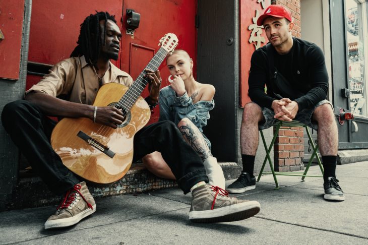Band aus drei Mitgliedern sitzt mit Instrumenten vor einer Wand