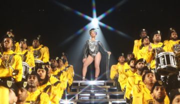 Beyoncé steht auf einer Bühnentreppe in einem glitzernden Einteiler und einem Gehstock, um sie herum eine Kapelle aus People of Colour